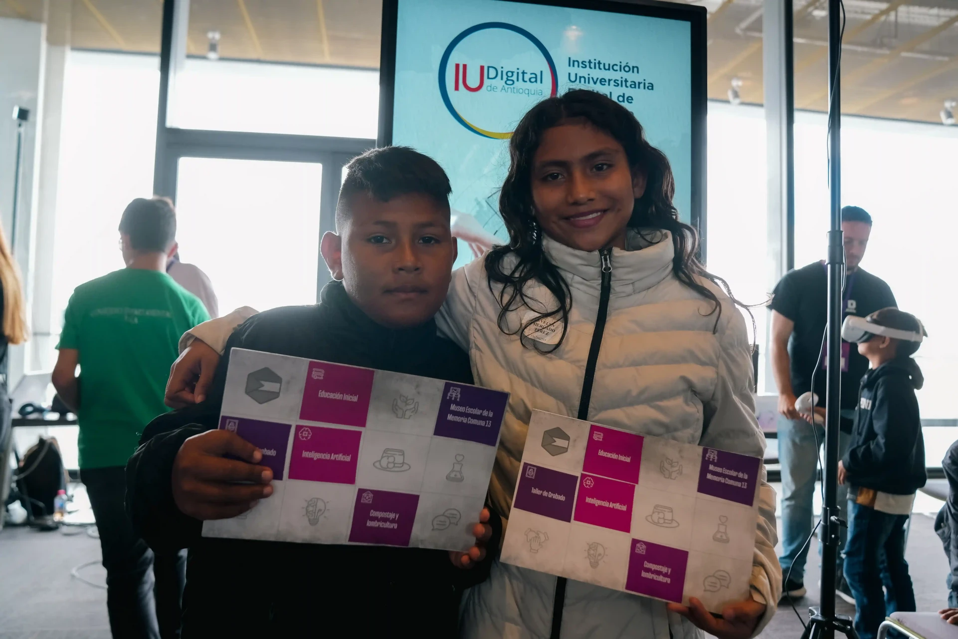 Dos estudiantes sostienen materiales educativos frente a la pantalla institucional de la IU Digital de Antioquia en el Foro Nacional de Educación 2025.