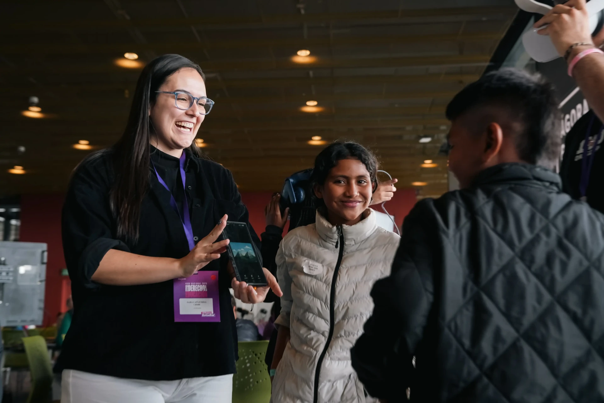 Participante del Foro Nacional de Educación 2025 muestra contenido en un dispositivo móvil mientras estudiantes exploran experiencias de realidad virtual.